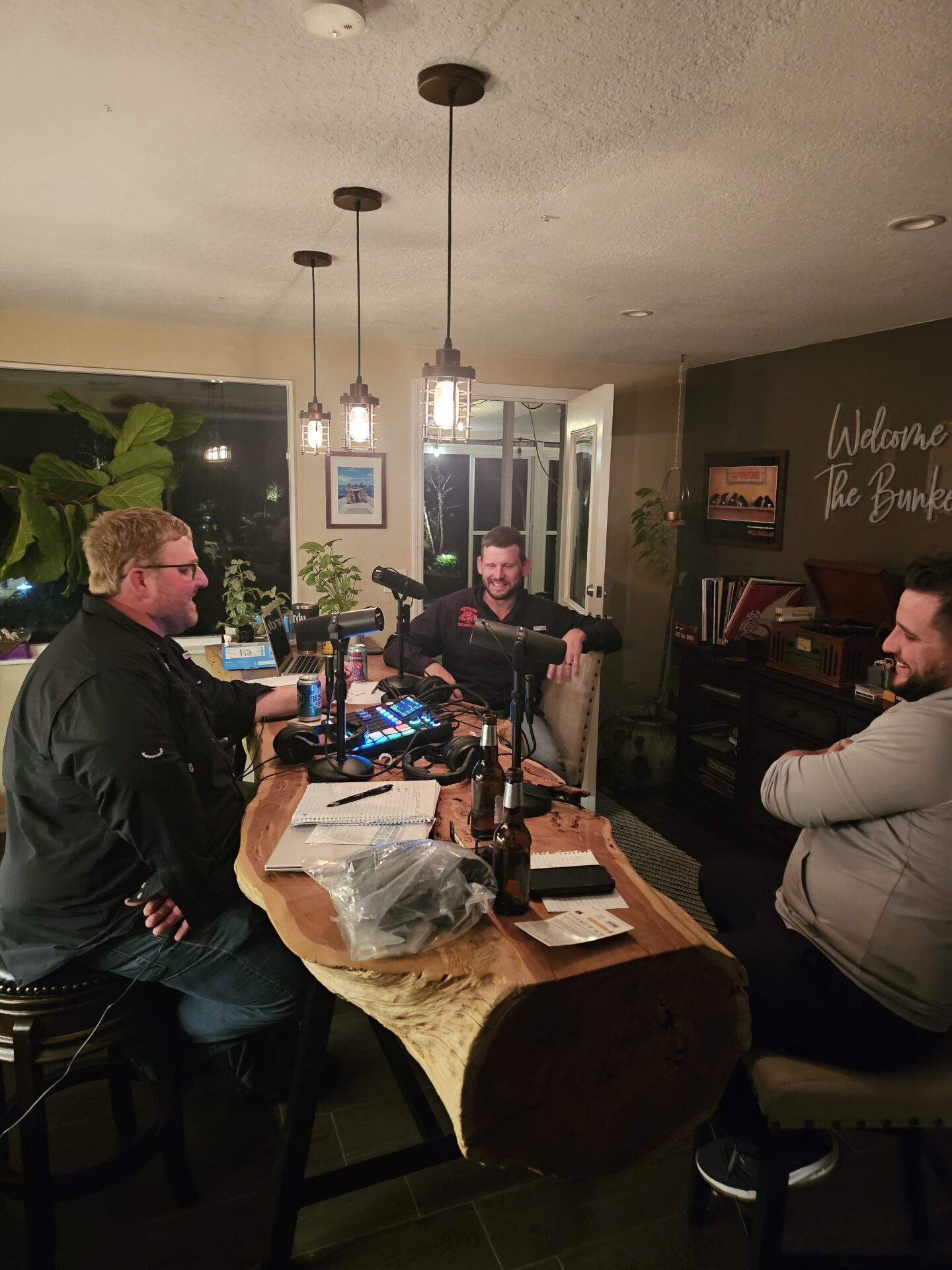 Three people sit around a wooden table with podcast equipment, beer bottles, and notebooks in a cozy room with warm lighting and plants.