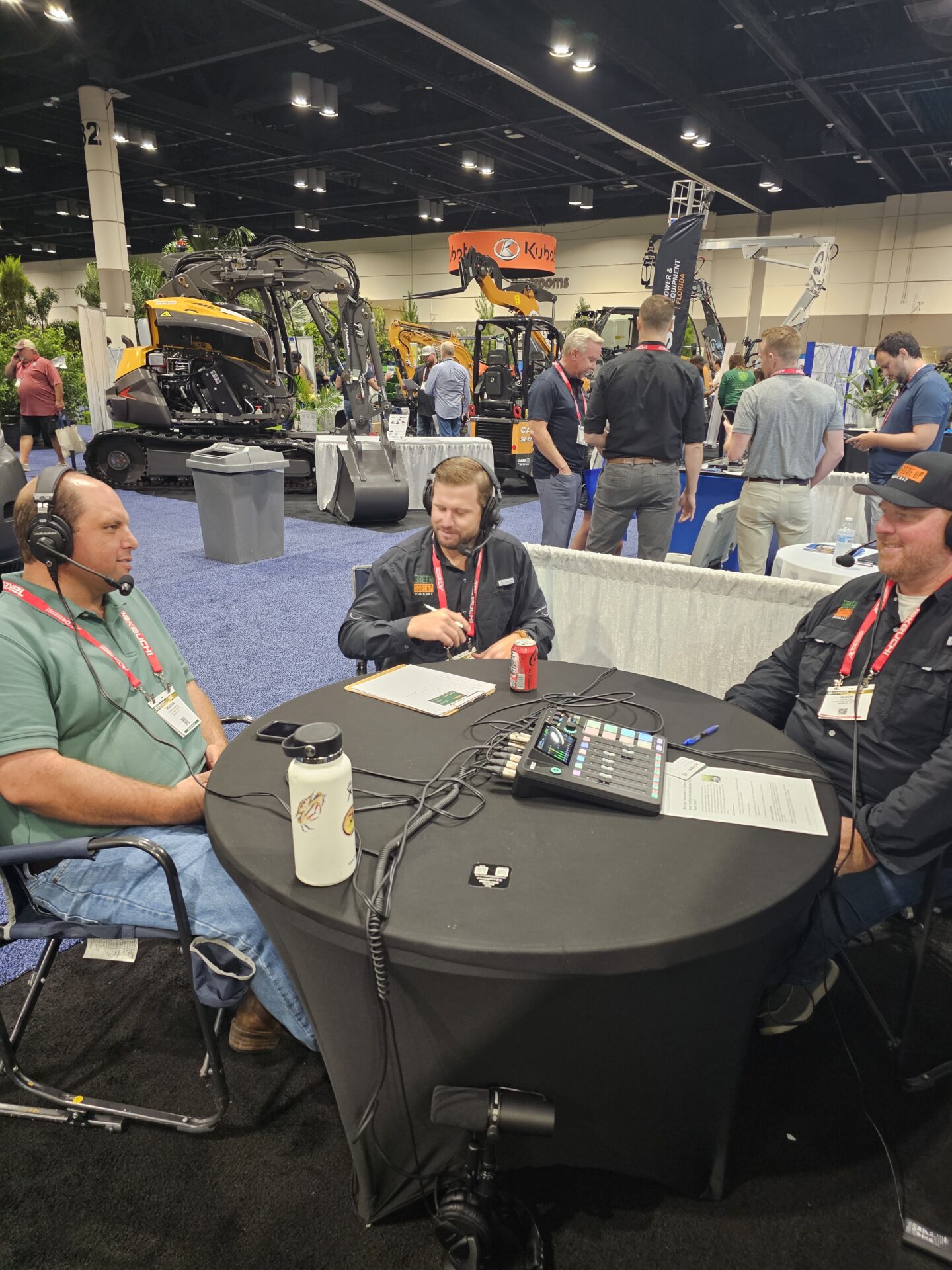 Three people sit around a table with recording equipment at an event. Construction machinery is displayed in the background.