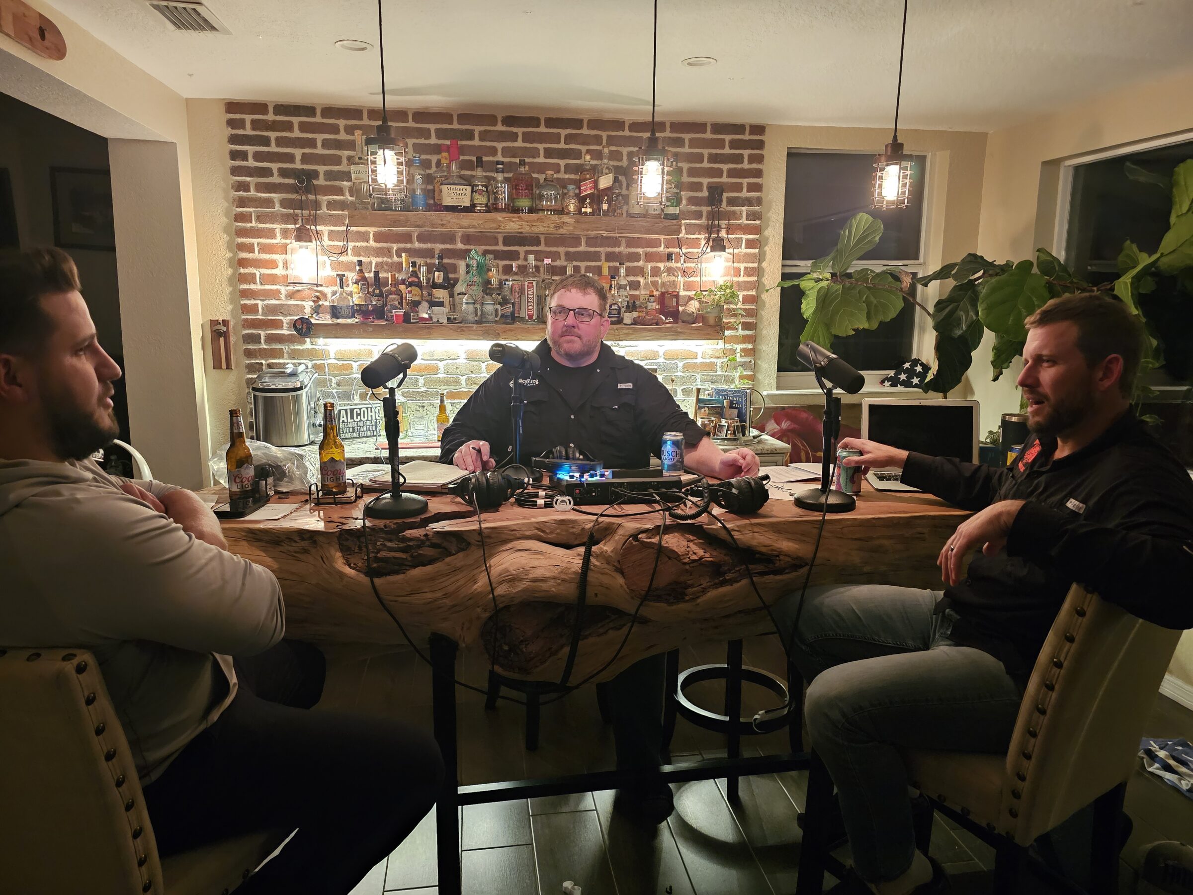 Three people recording a podcast at a wood table with microphones, bottles, and audio equipment. Brick wall and shelves with bottles in the background.