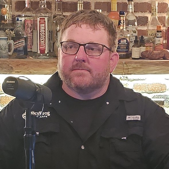 A person with glasses and a beard is in a bar setting with various bottles and a microphone in front of them.