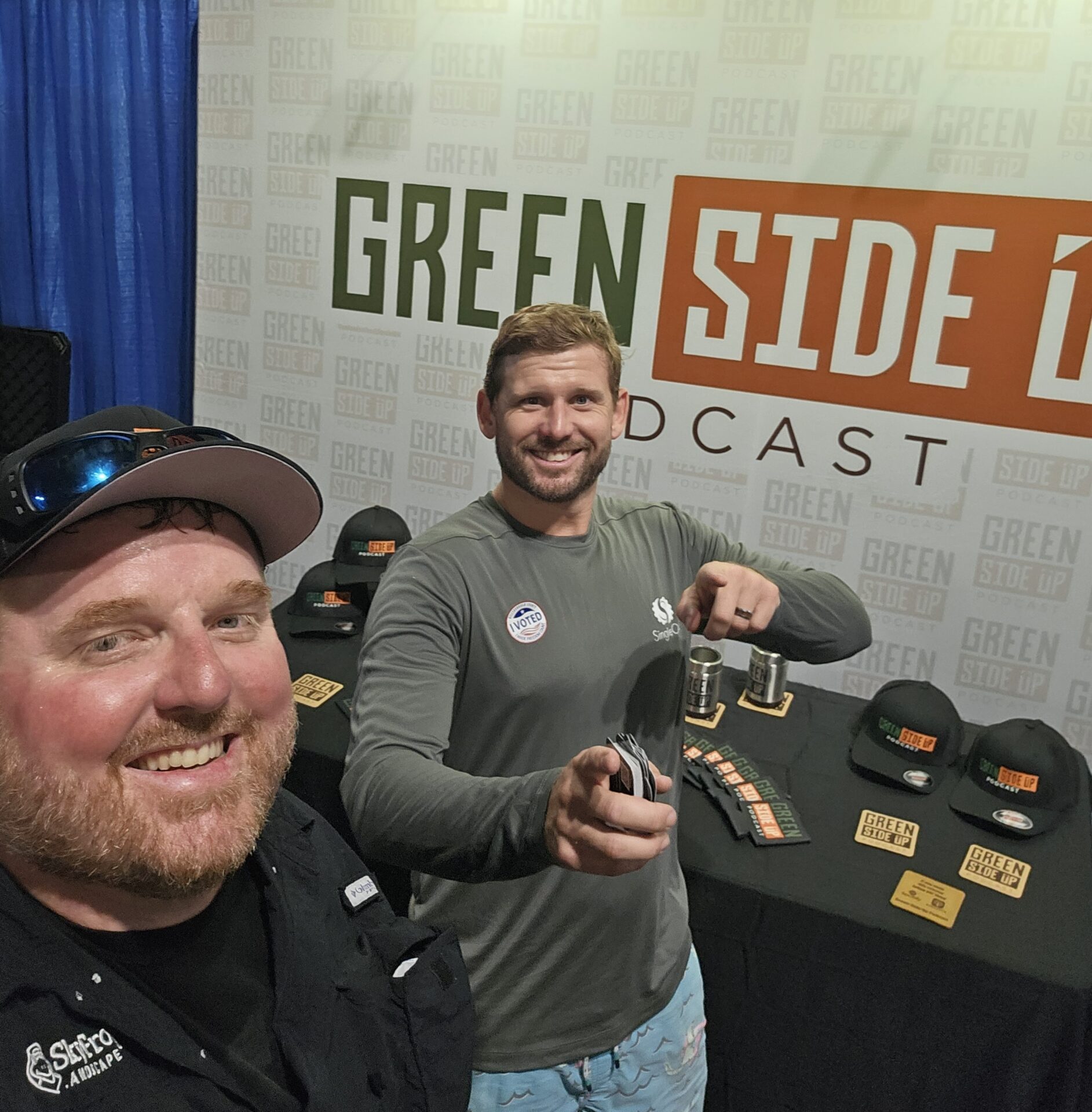 Two people smiling in front of a Green Side Up podcast booth, with merchandise like hats and cards displayed on a table.