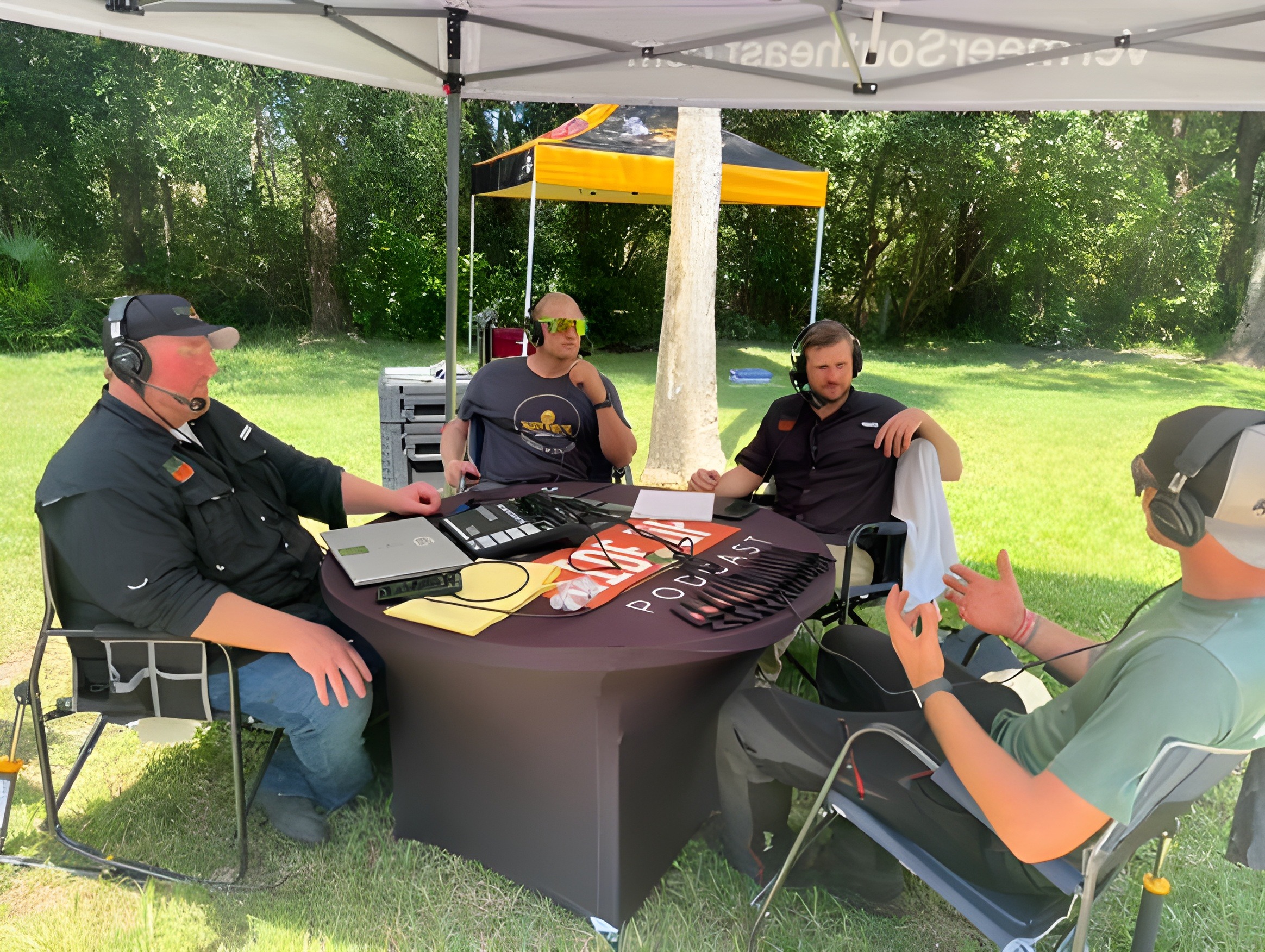 Four people wearing headsets engaged in a podcast discussion under a tent in a grassy outdoor setting, with recording equipment on a table.